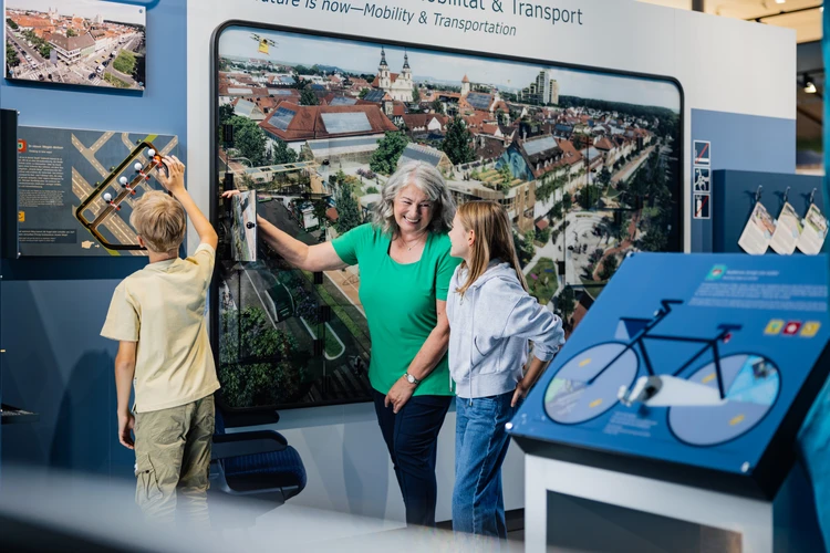 Gro&szlig;mutter mit zwei Kindern vor einem Bild mit Zukunftsvision f&uuml;r Mobilit&auml;t.